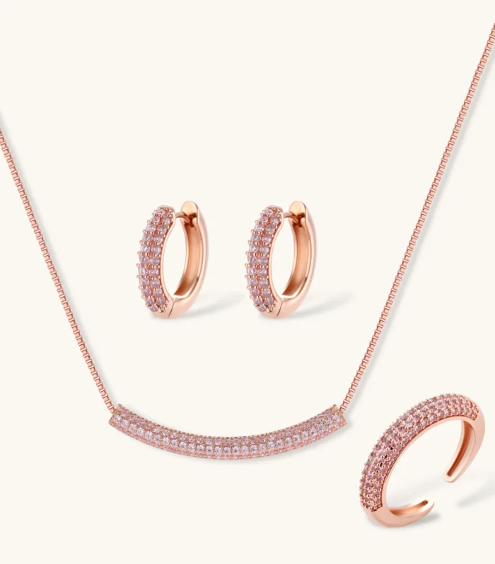 Rose gold jewelry set with pavé cubic zirconia stones including curved necklace, hoop earrings, and adjustable ring on cream background.
