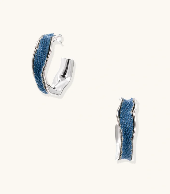 Silver huggie hoops with blue denim fabric inlay strip and polished white gold plated finish on cream background.