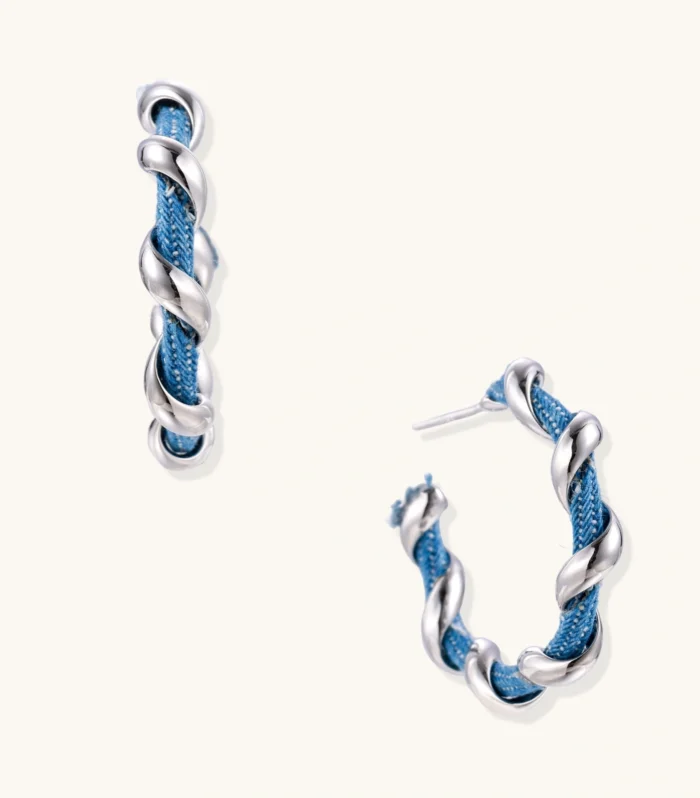 White gold-plated hoops intertwined with blue denim fabric in a twisted rope design on cream background.