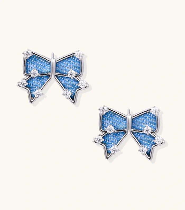 Blue denim bow stud earrings with white gold plating and crystal accents on cream background