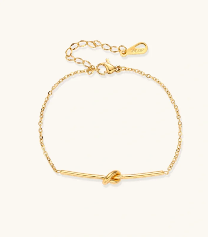 Delicate gold chain bracelet with minimalist bar and centered knot detail on cream background