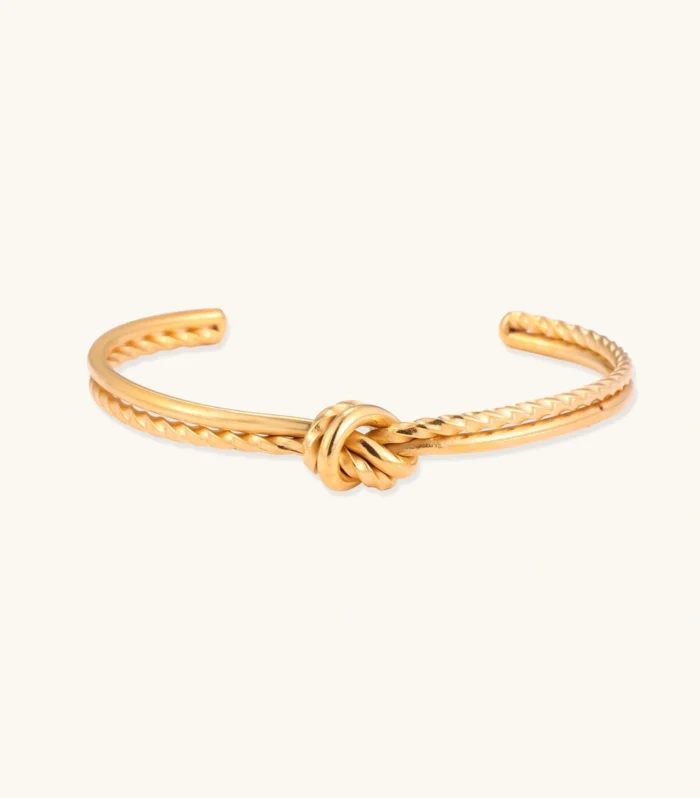 Gold cuff bracelet with twisted rope texture and decorative knot centerpiece on cream background.
