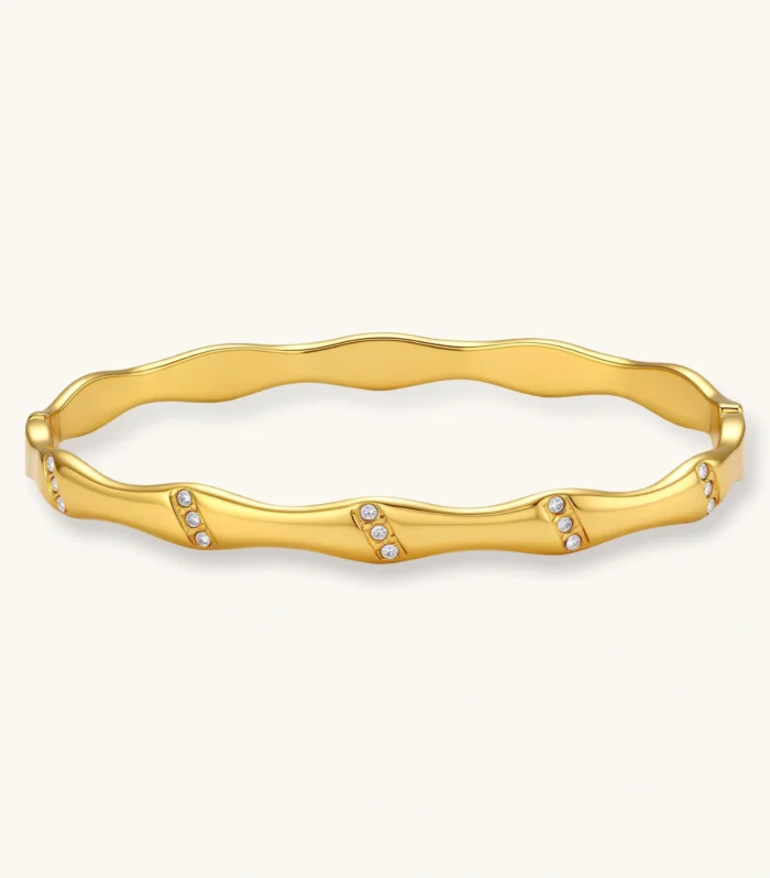 Gold-toned wavy bangle with bamboo-style segments and CZ crystal clusters on cream background. 18K plated stainless steel.
